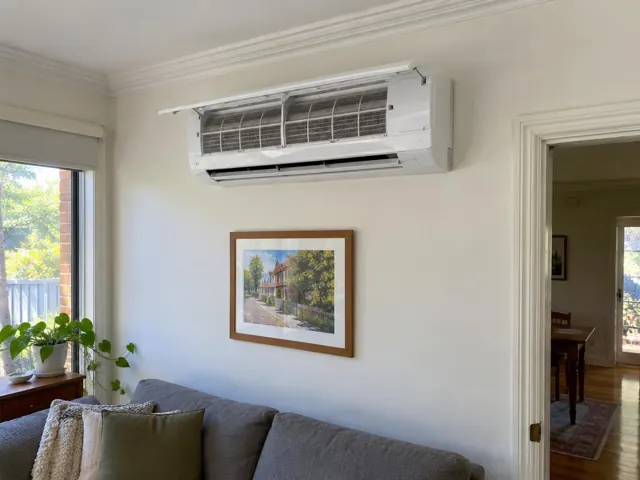 Clean split system with open front panel and visible filters on a white wall above a sofa in a living room in Mile End
