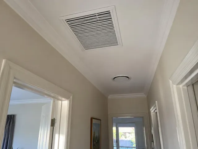 Ceiling return-air grille and round wall vent in a hallway with white trim in Plympton