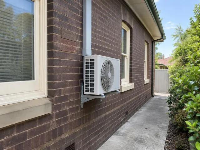 Outdoor air conditioner unit mounted on a brick house wall beside a window in Plympton
